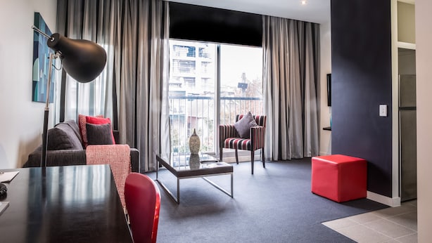 A modern living room with a sofa, armchair, and ottoman, a glass coffee table, and a large window with curtains. - Adara Hotel Richmond (Richmond)