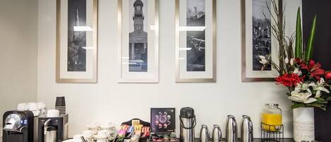 Zona de recepción de hotel con cuadros enmarcados en la pared, estación de café con diversos utensilios para prepararlo, y un jarrón con flores.