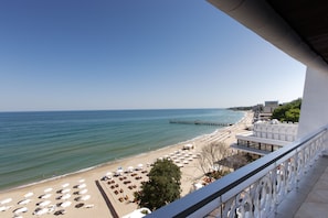 A beach view from a balcony with sun loungers and umbrellas, a pier extending into the sea, and buildings in the background.