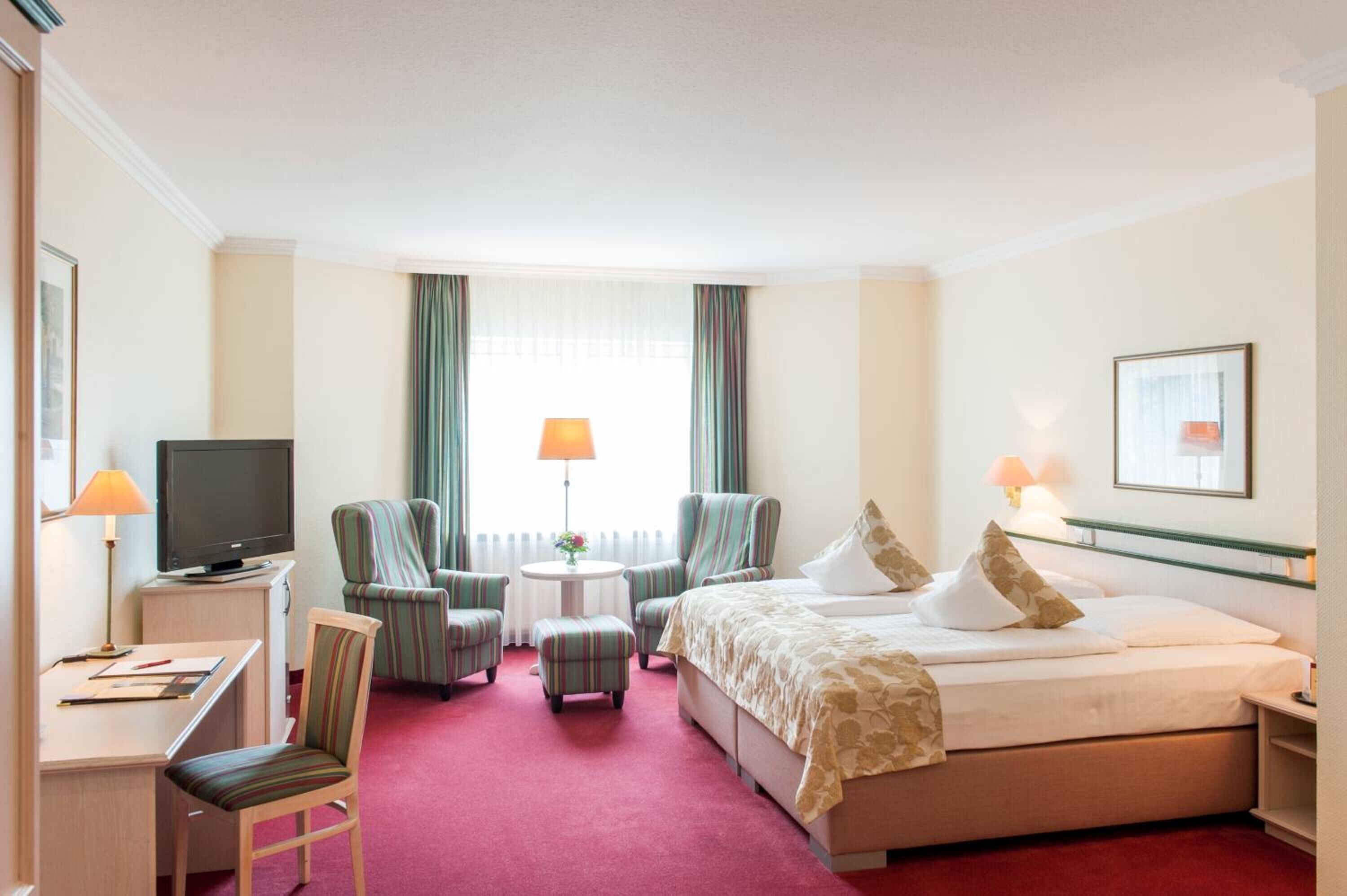 Habitación de hotel con cama, dos sillones, un escritorio con computadora, televisor y una mesita de noche con lámpara.