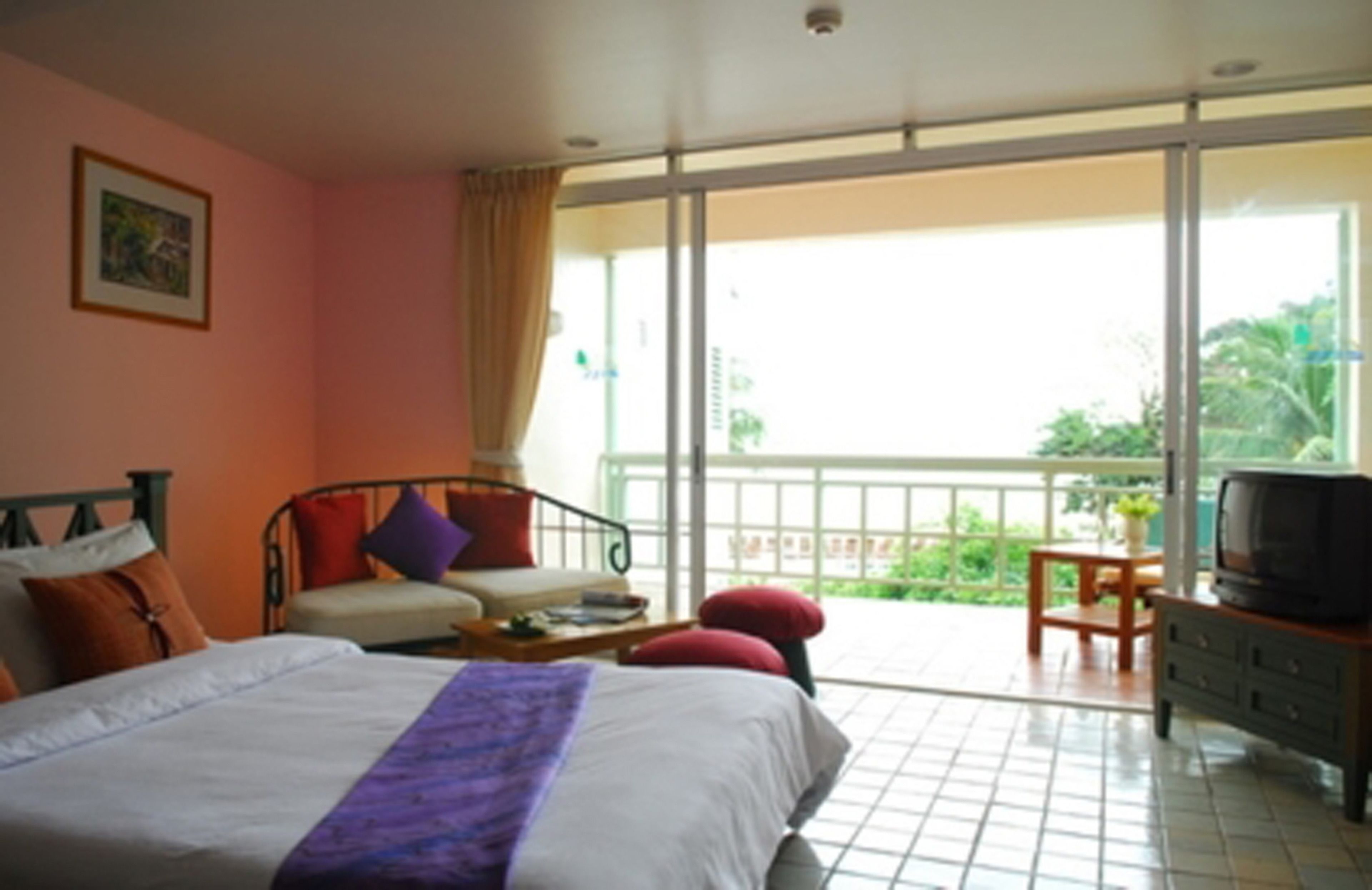 Habitación de hotel con cama, sofá, televisión y balcón con vista a follaje.