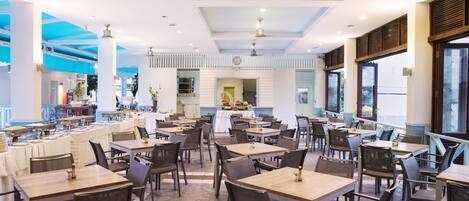 A spacious restaurant with multiple tables and chairs, a central serving area, and large windows allowing natural light.