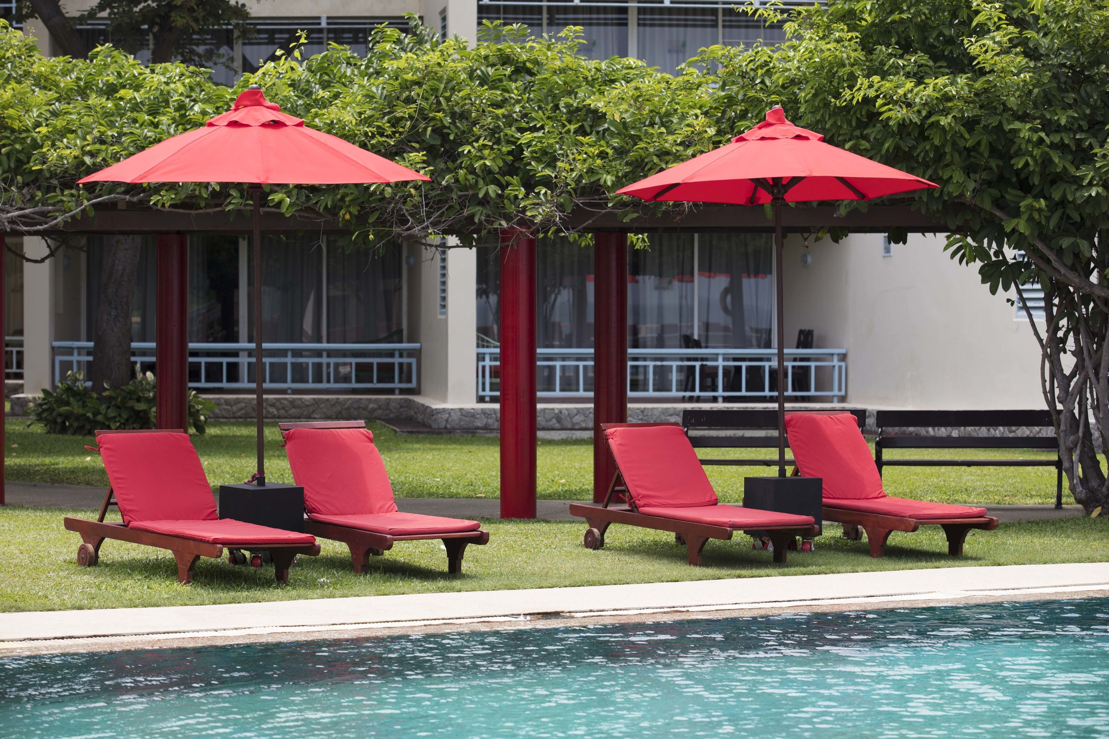 A poolside area with red lounge chairs and umbrellas, a clear pool, and a building with large windows in the background.