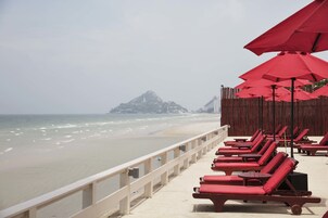 Vista de la playa con sillas de reposo rojas y sombrillas, una valla de madera y una montaña a lo lejos.