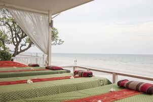 Varanda com duas camas, cada uma com colcha estampada em verde e almofadas vermelhas, com vista para uma praia e o mar.