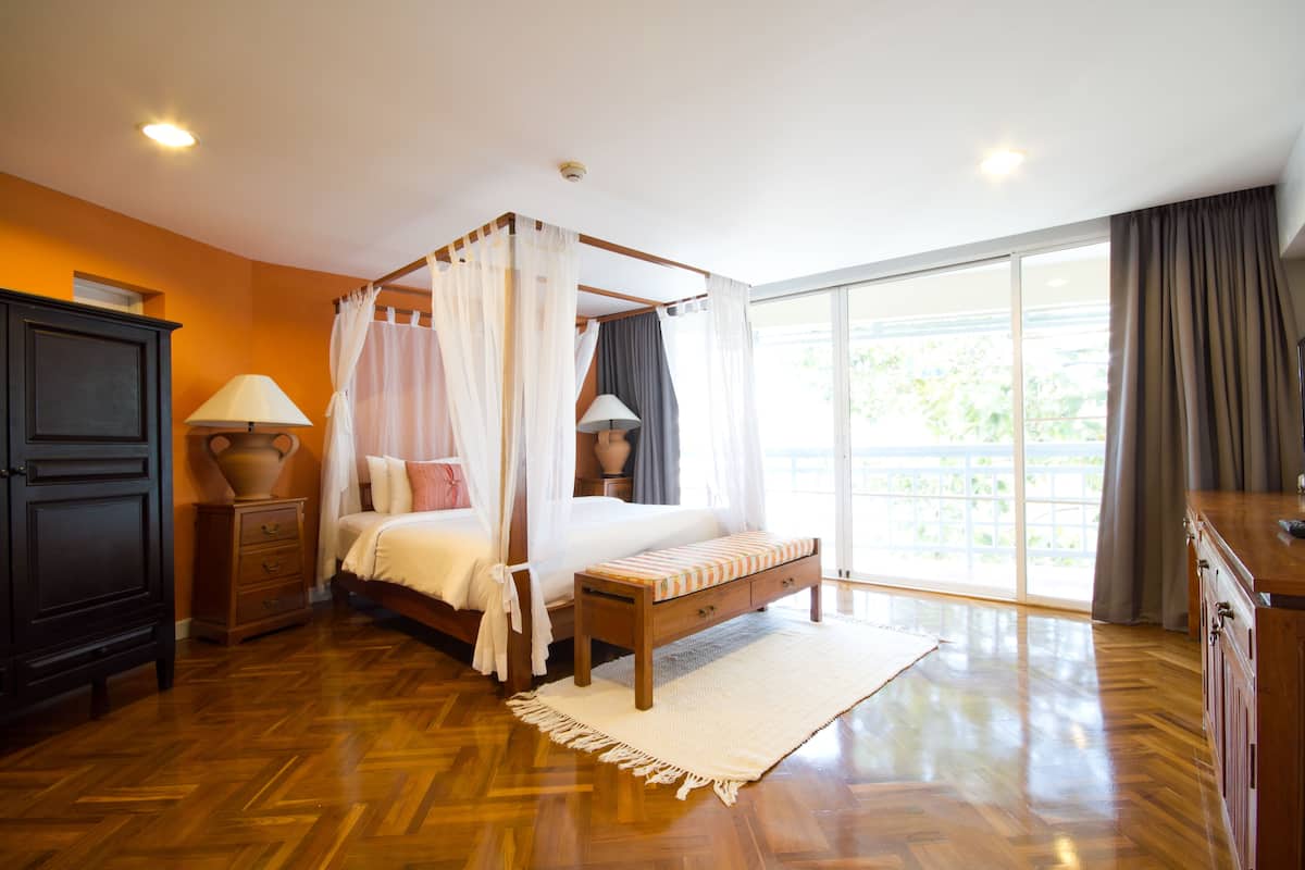 A four-poster bed with a canopy, a wooden bench, a nightstand with a lamp, and a large window with curtains.
