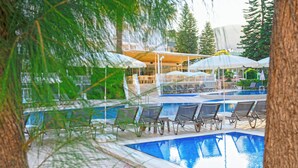 Área de piscina com espreguiçadeiras e guarda-sóis, cercada por árvores e um edifício com espaço de estar coberto.