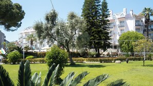 Um parque com grama, árvores e um edifício com varandas ao fundo.