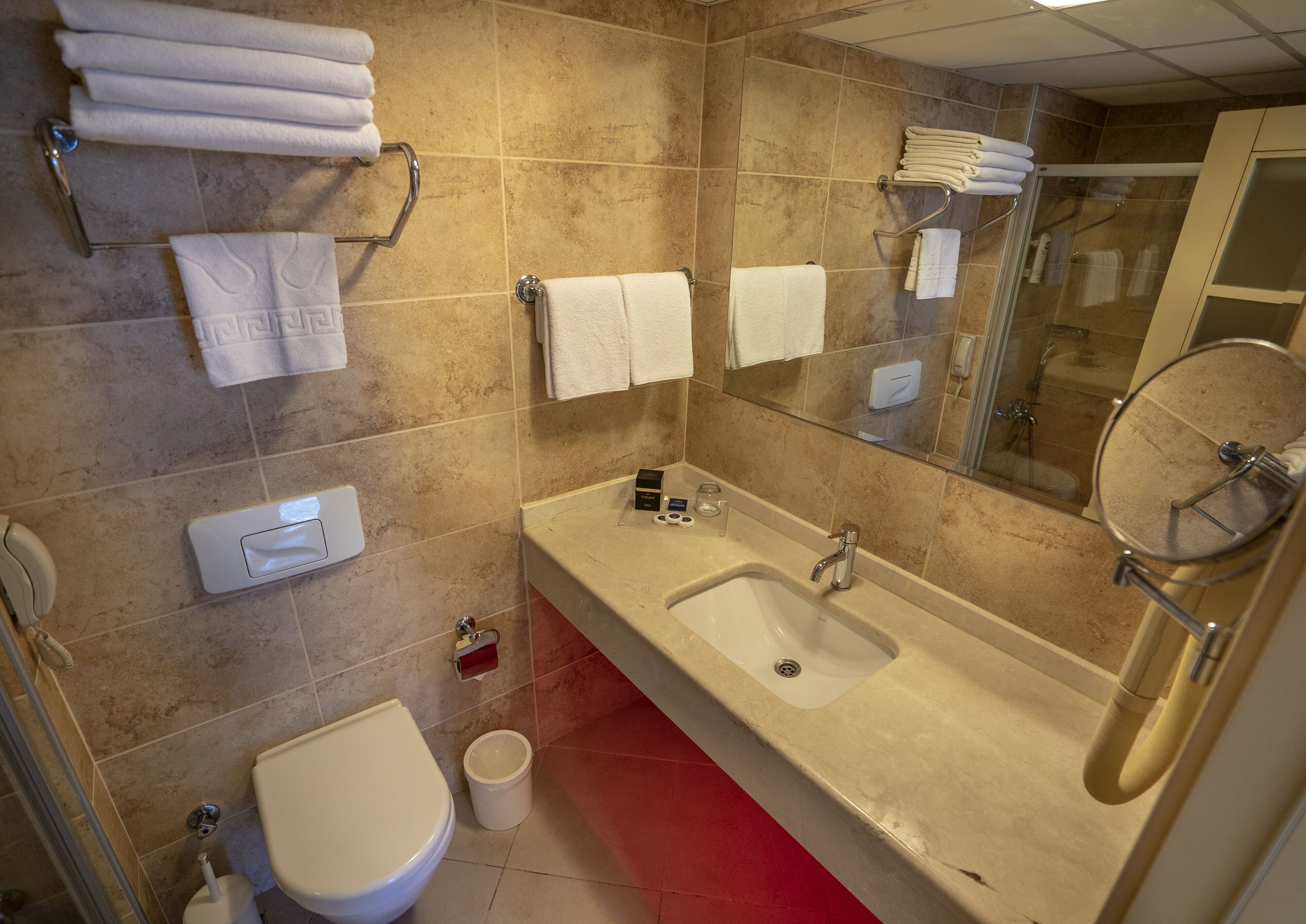 A bathroom with a sink, mirror, and toilet. There are towels hanging on hooks and a shower visible in the background.