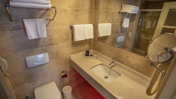 A bathroom with a sink, mirror, and toilet. There are towels hanging on hooks and a shower visible in the background.