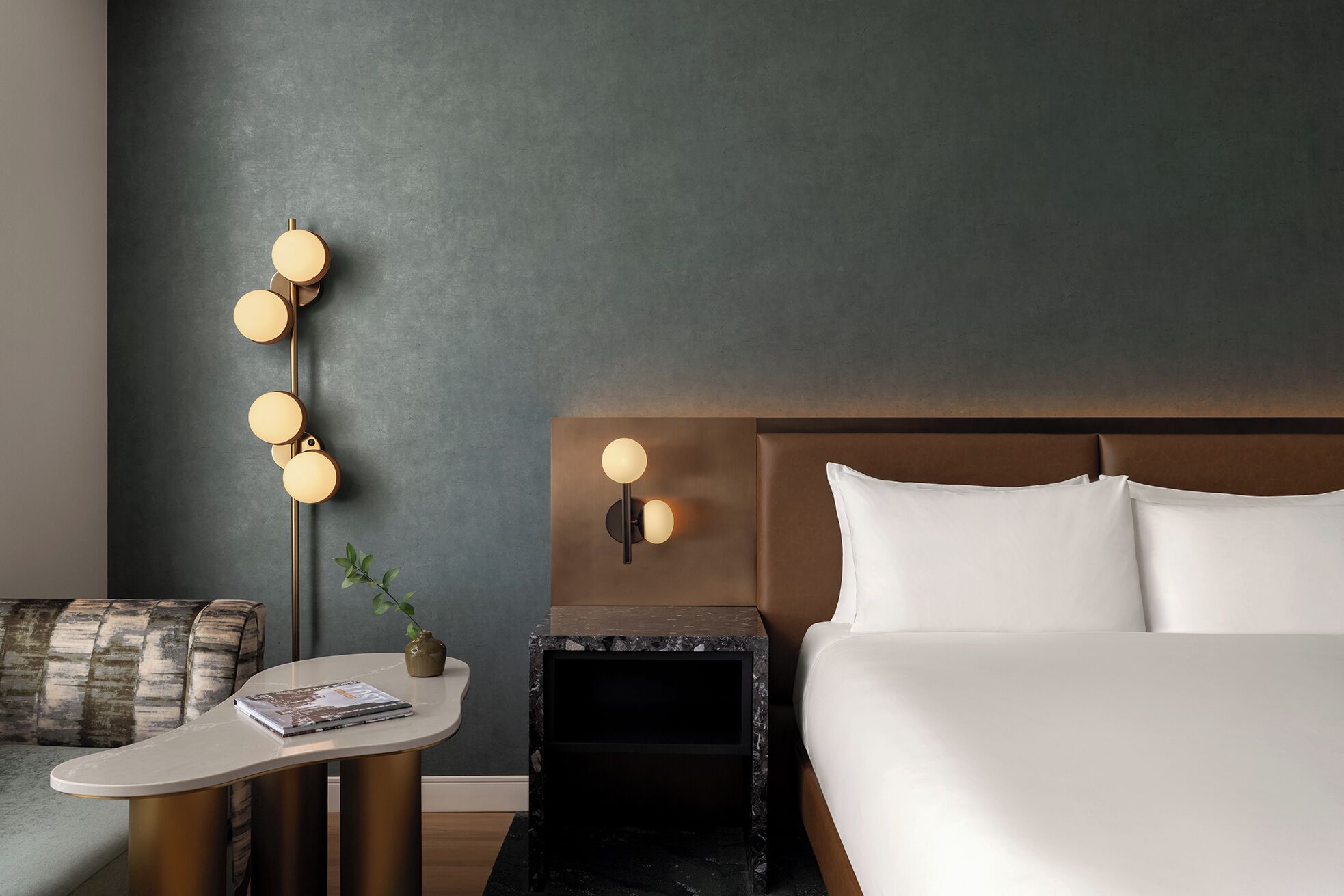 A modern hotel room with a bed, bedside table, lamp, and a small plant.