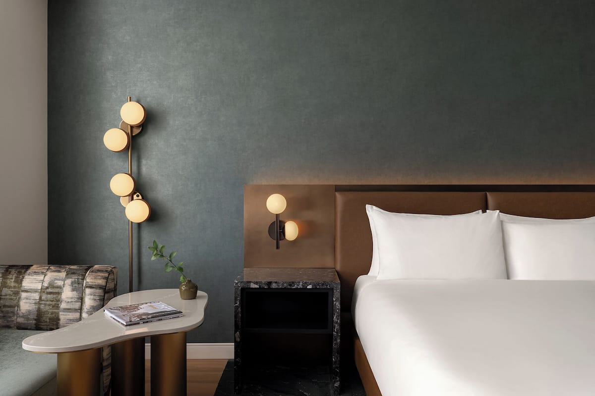 A modern hotel room with a bed, bedside table, lamp, and a small plant.