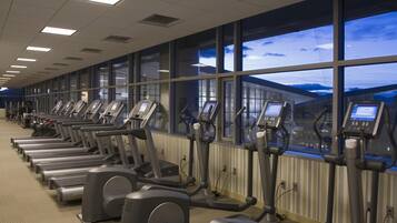 A row of stationary bikes in a gym with large windows and a view of a building outside.