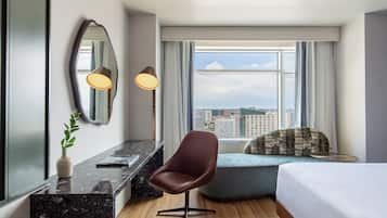 A modern hotel room with a large bed, a desk with a marble top, a brown leather chair, a round table, a large window with a city view, and a mirror with two lamps.