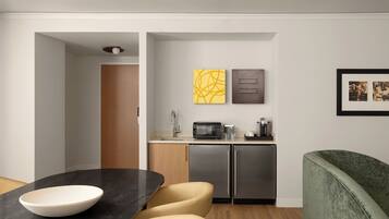 A modern kitchen with a microwave, coffee maker, and refrigerator. There is a dining area with a bowl on a table and chairs.
