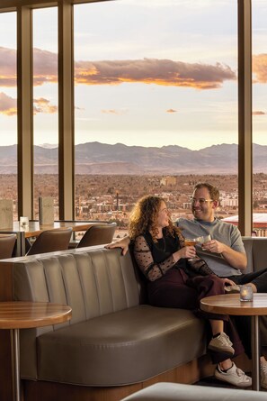 A couple seated on a leather couch with a cityscape view through large windows. - Hyatt Regency Denver at Colorado Convention Center (Denver)