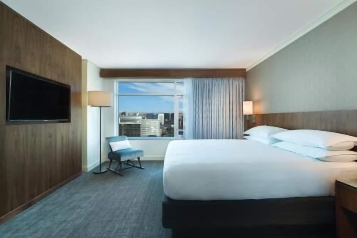 Habitación de hotel con cama, televisor montado en la pared, una silla y ventana con vista a la ciudad.
