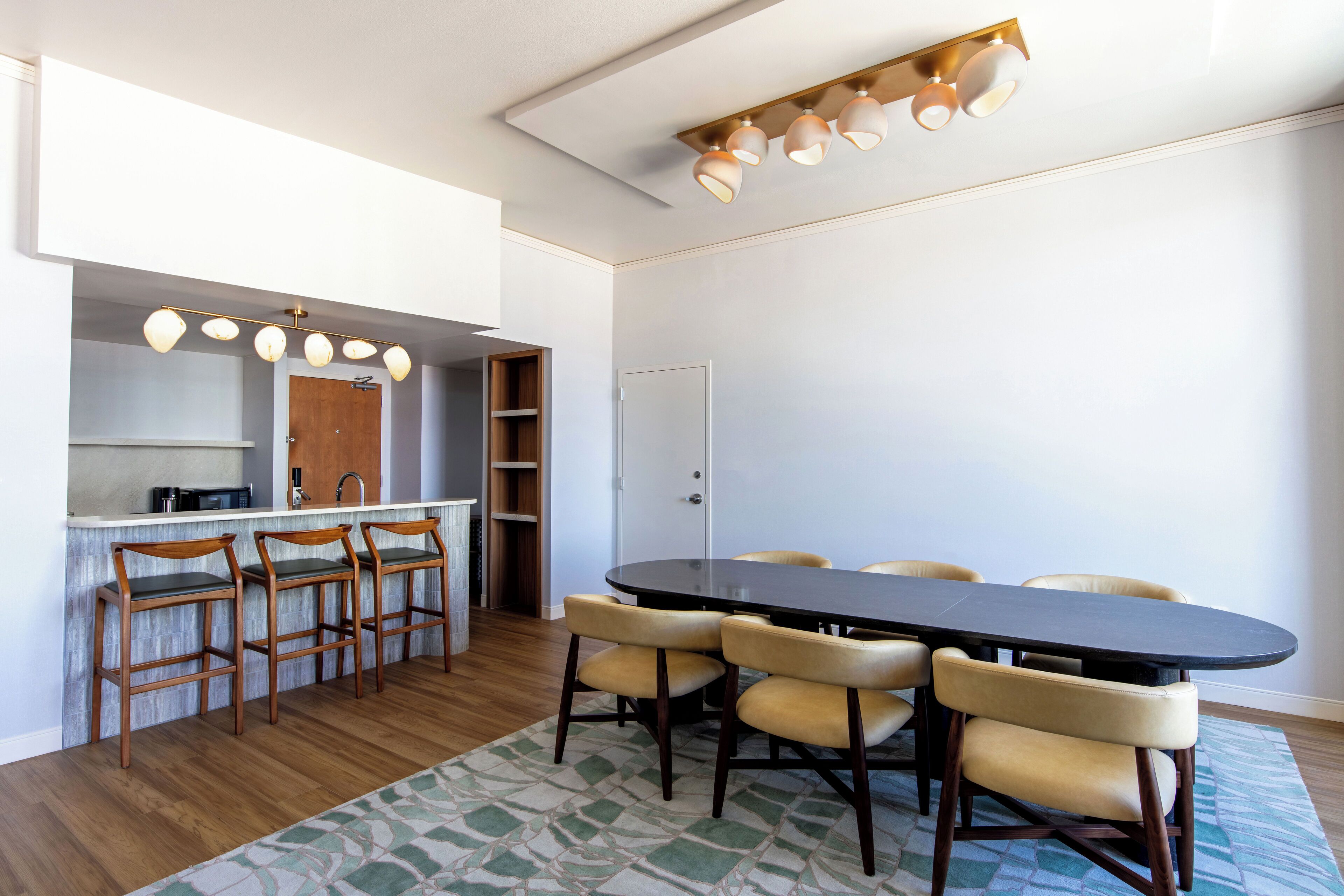 A dining area with a long table and six chairs, a bar area with stools, and a kitchen visible through an open door.