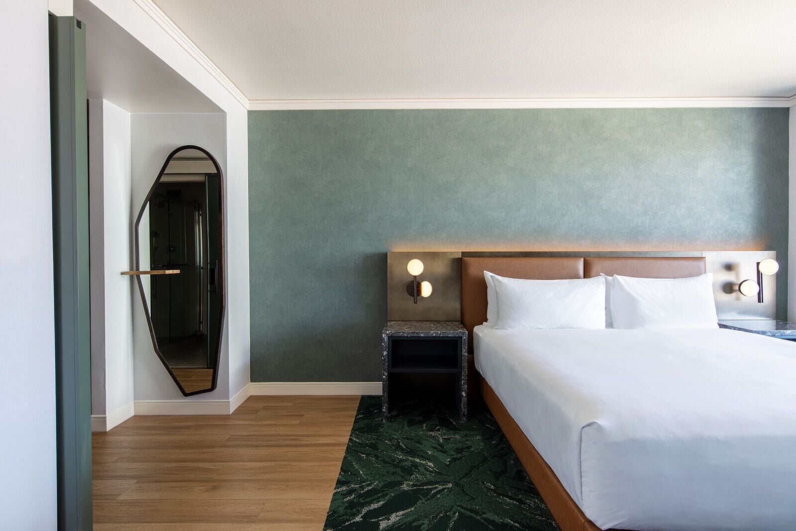 A hotel room with a bed, a nightstand, a mirror, and a green accent wall.