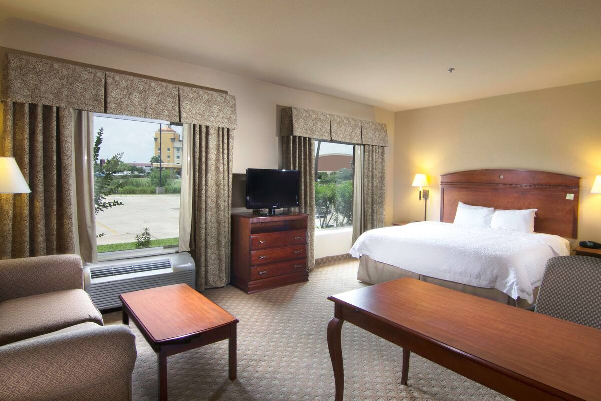 A hotel room with a bed, a desk, a chair, a dresser, a television, and large windows with curtains.