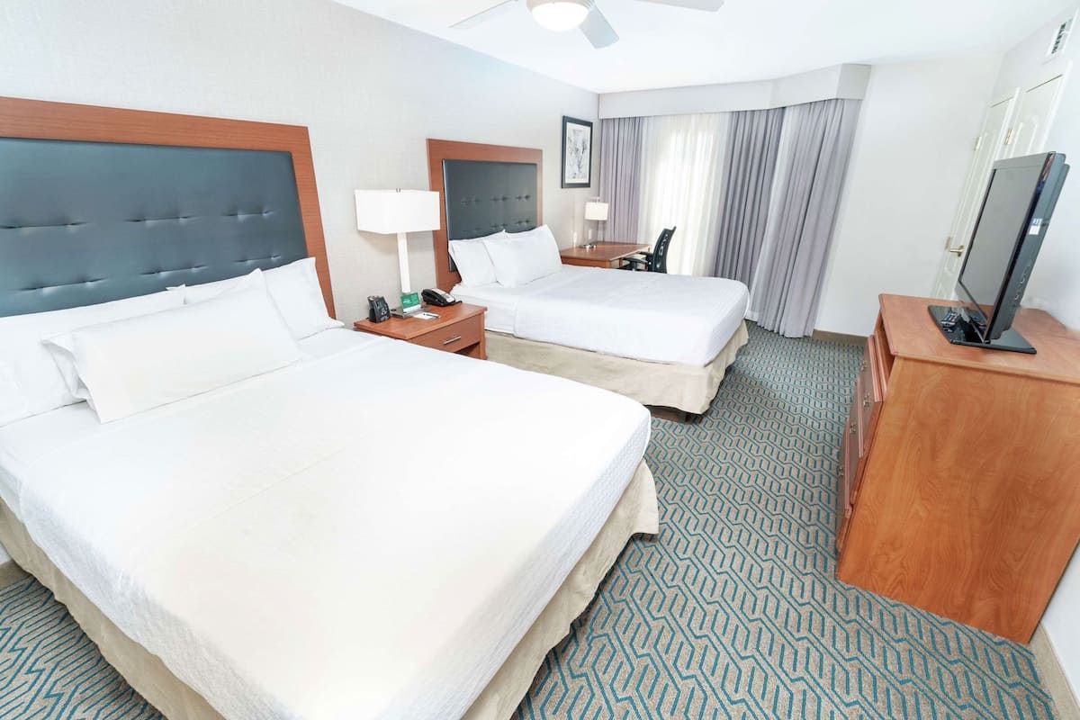 A hotel room with two beds, a desk, a chair, a TV, and a ceiling fan.