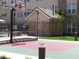 Una cancha de baloncesto con aro, un bote de basura y un edificio con fachada de piedra y ventanas.
