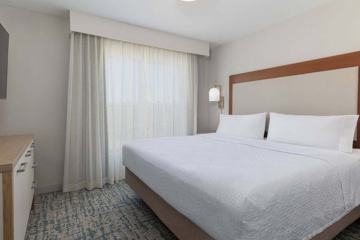 Een hotelkamer met een groot bed, een houten hoofdbord, een nachtkastje en een raam met dunne gordijnen.