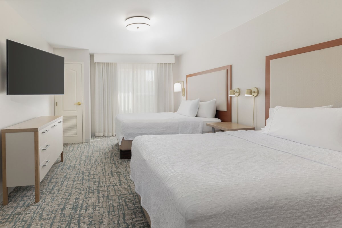 A hotel room with two beds, a flat-screen TV, a desk, and a window with curtains.