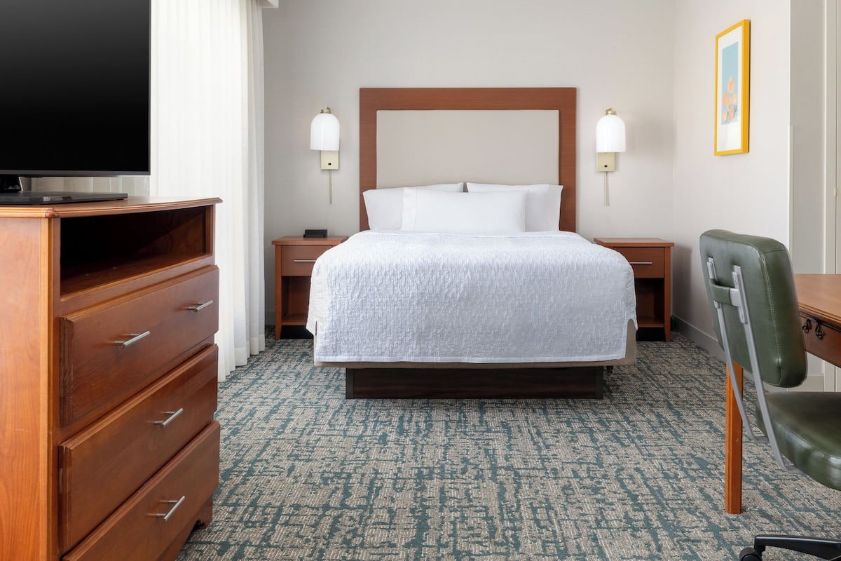 A hotel room with a large bed, wooden dresser, two bedside tables, a desk with a chair, and a flat-screen TV.