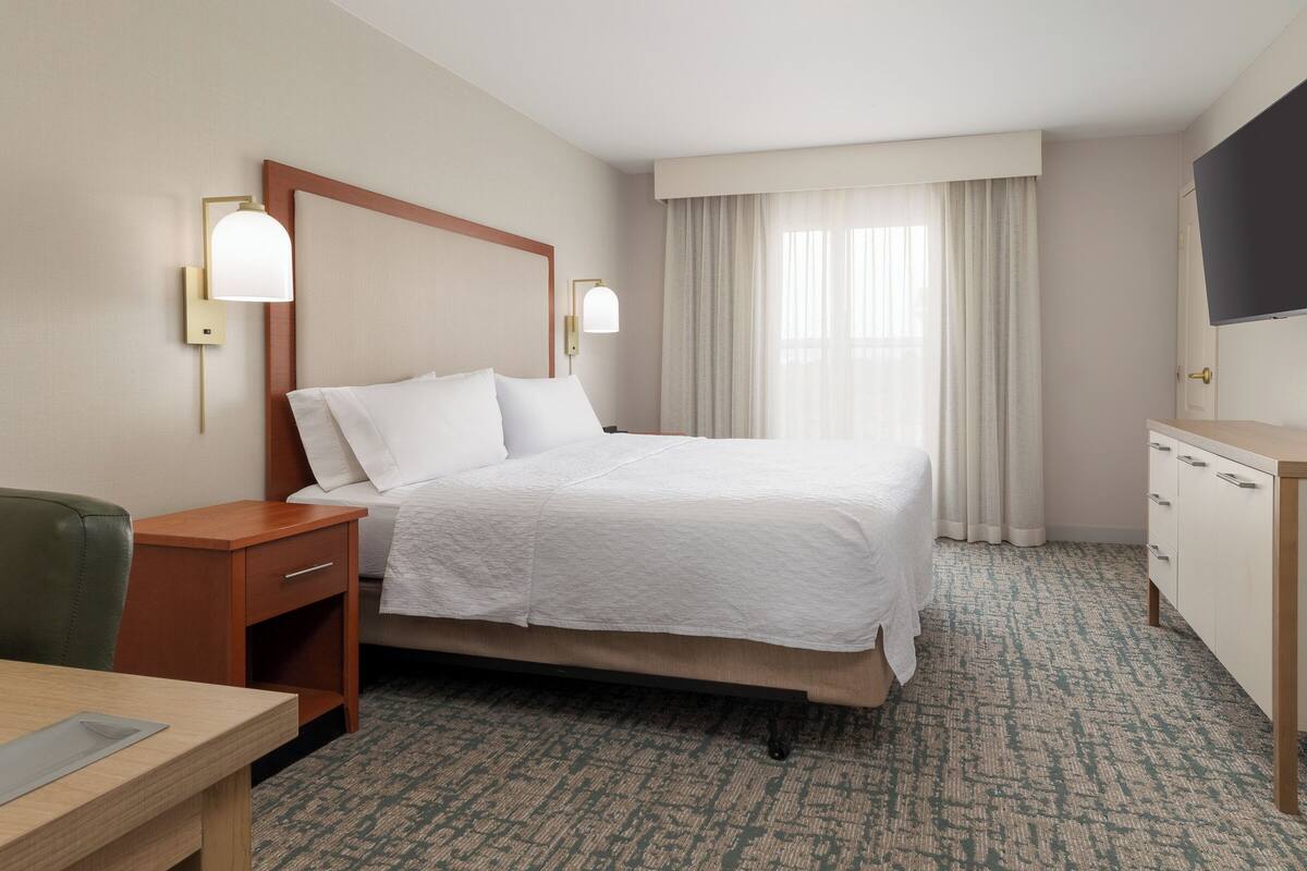 A hotel room with a bed, bedside tables, a desk, a chair, a TV, and a window with curtains.