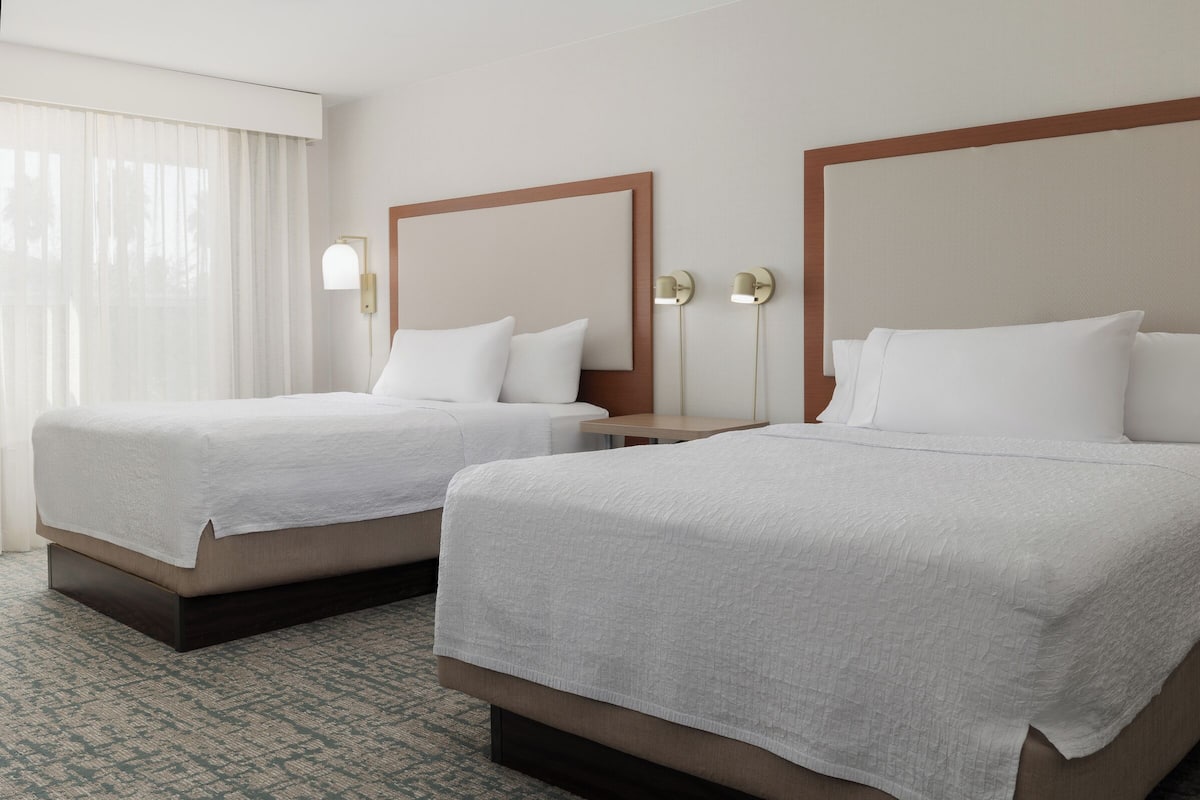 Two double beds with white linens, a wooden headboard, a nightstand, and a wall-mounted lamp.