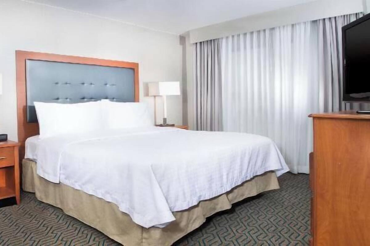 Een hotelkamer met een bed, twee bedlampen, een televisie, een bureau met stoel, en een raam met gordijnen.