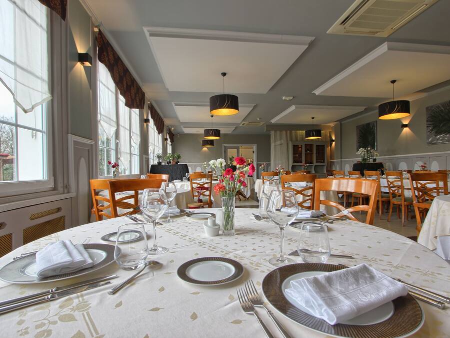 Comedor con mesas redondas dispuestas para cuatro personas, cada una con mantel blanco, platos, cubiertos y un florero con flores.