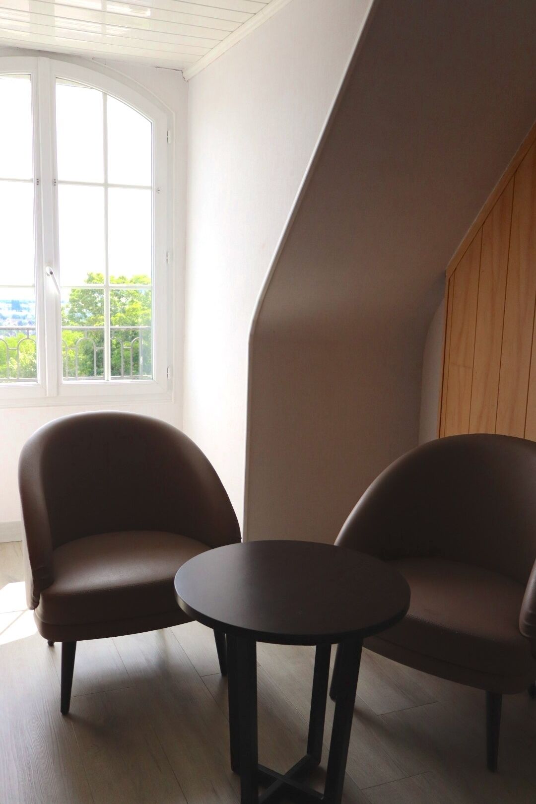 Une pièce avec deux fauteuils marron et une petite table ronde, une porte blanche donnant sur un espace verdoyant, et un plafond en pente.