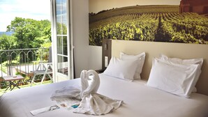 A hotel room with a bed, white pillows, a towel art, a balcony with a view of greenery and a building, and a large wall painting of a field.