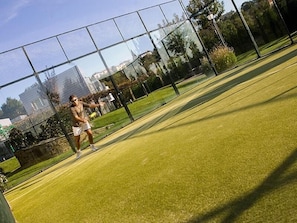 Tenniskenttä, jossa on verkko, pelaaja toiminnan keskipisteessä ja kentän ympärillä oleva aita.