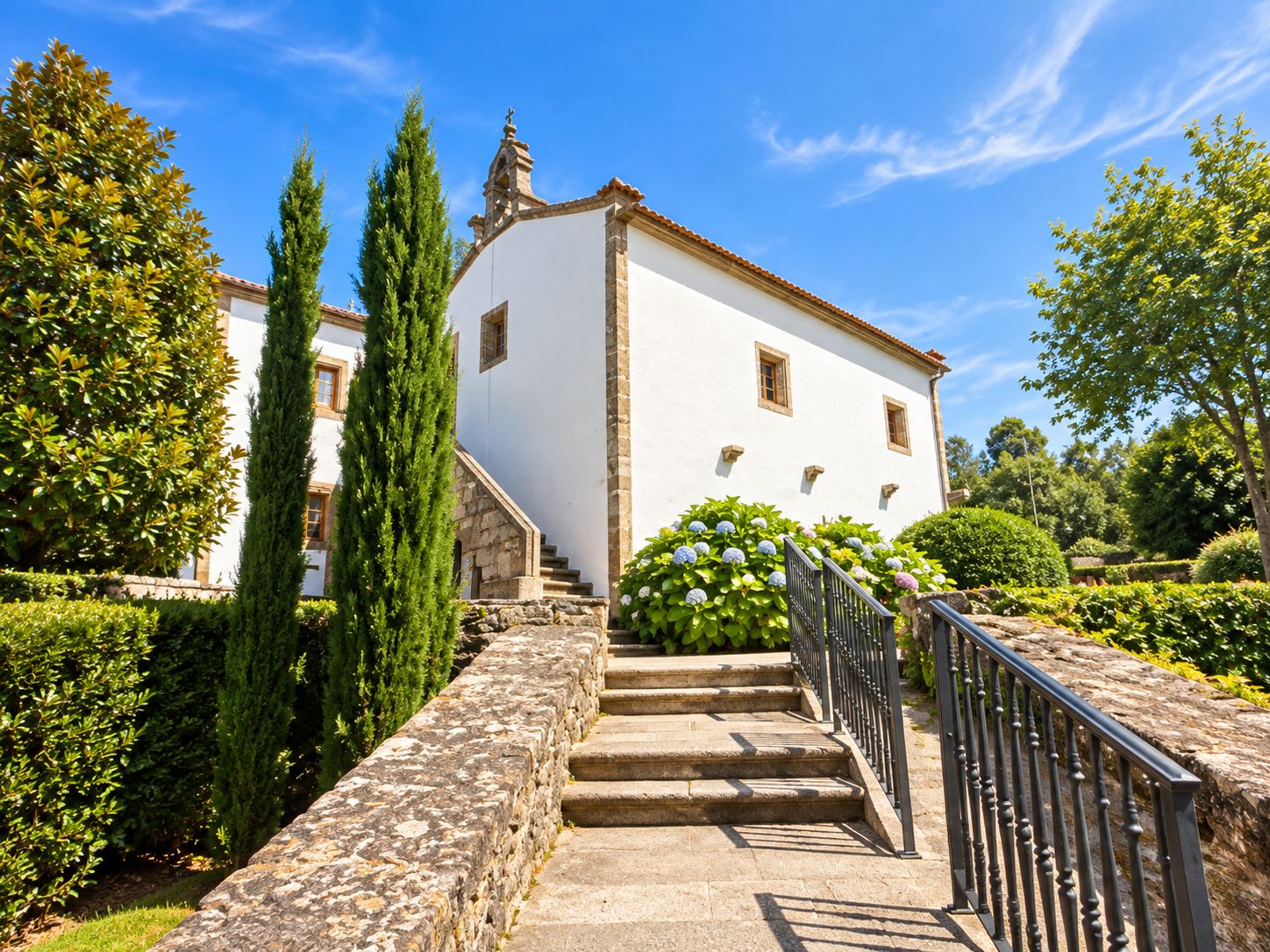 Un edificio blanco con una escalera de piedra que lleva a una puerta, rodeado de follaje y árboles.