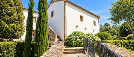 Un edificio blanco con una escalera de piedra que lleva a una puerta, rodeado de follaje y árboles.