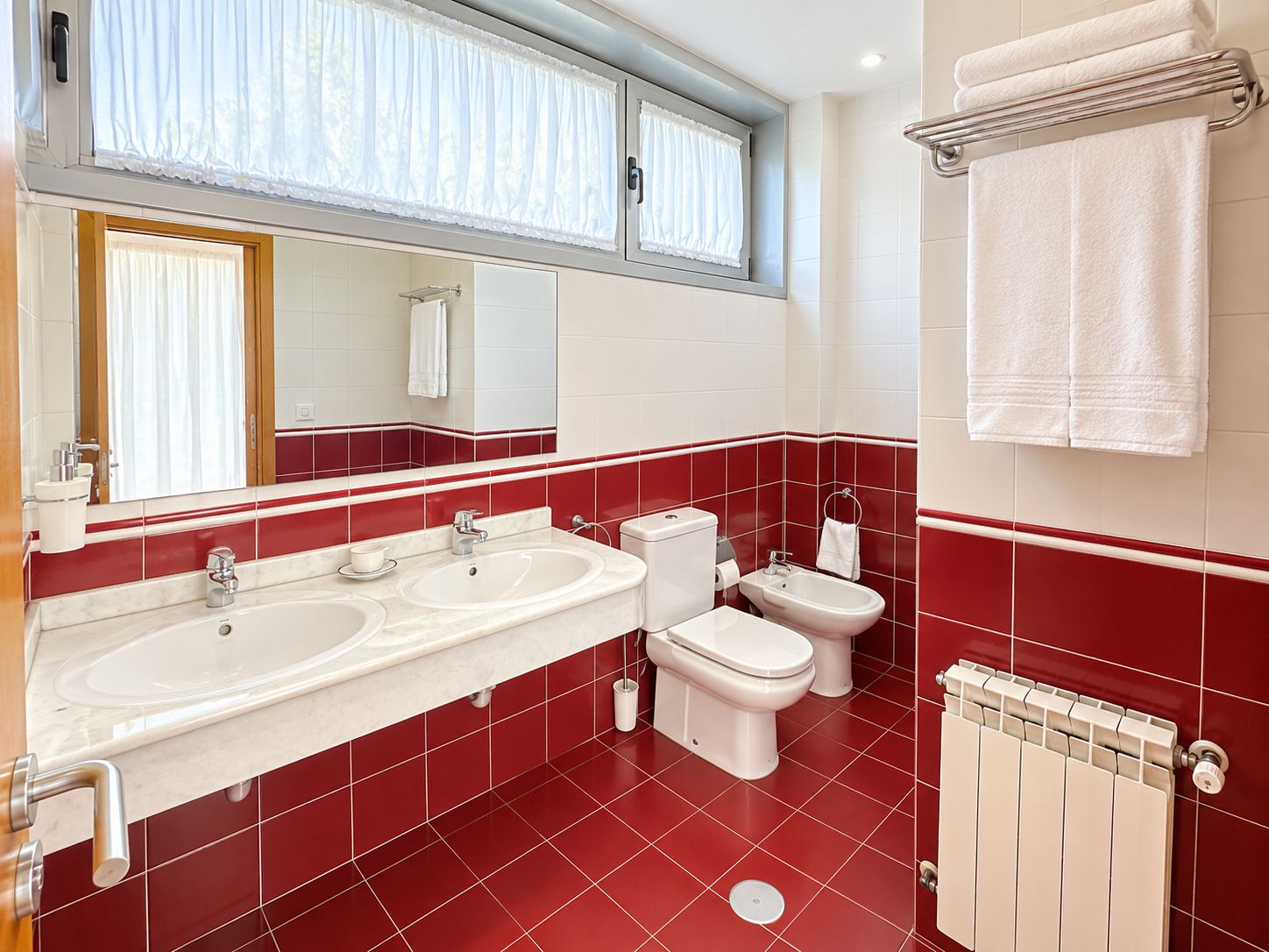 Een moderne badkamer met twee wastafels, toilet, bidet en een raam met gordijnen.