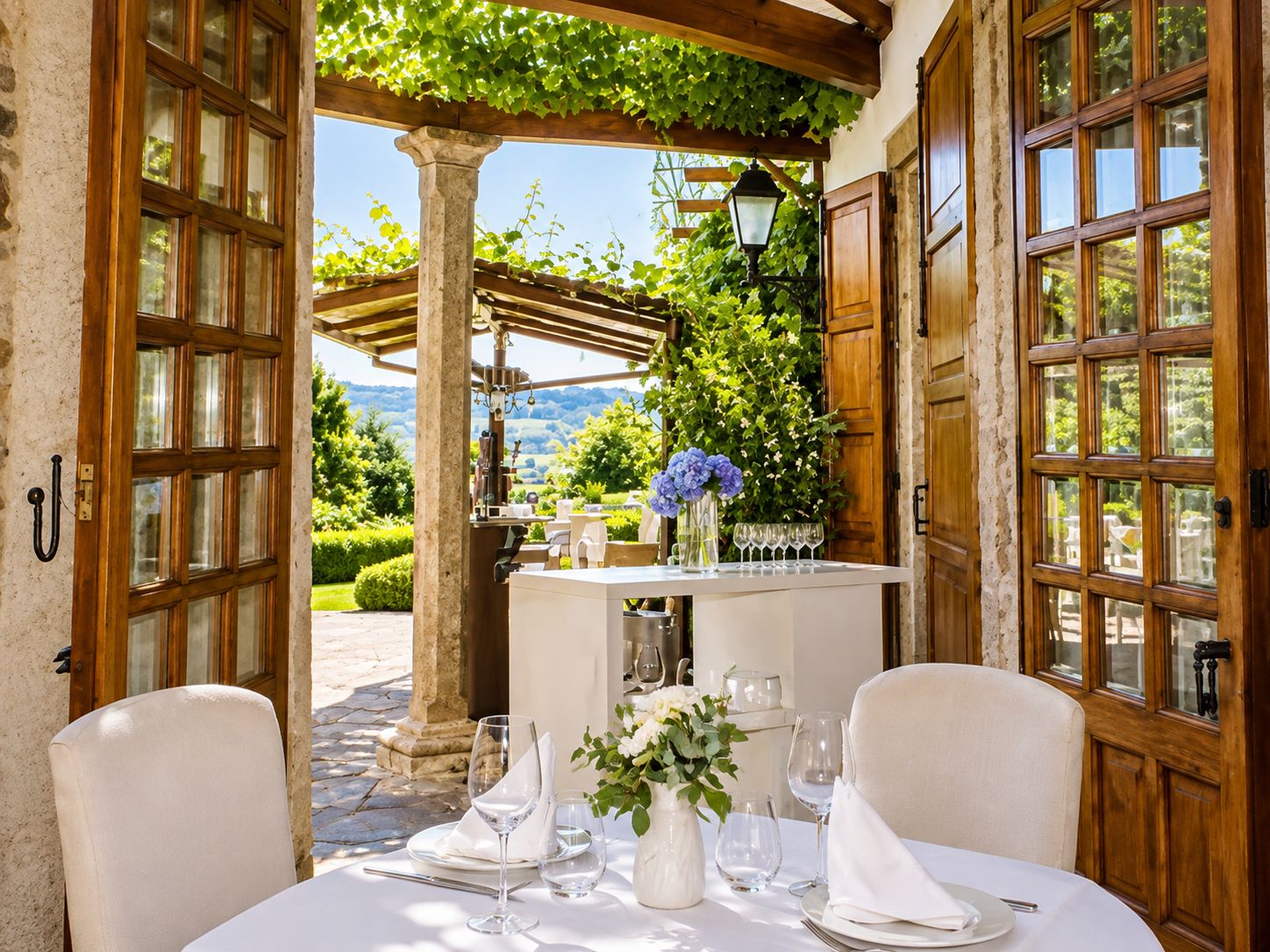 Un espacio de comedor al aire libre con una mesa dispuesta para dos, un pilar de piedra y una pérgola con vegetación.