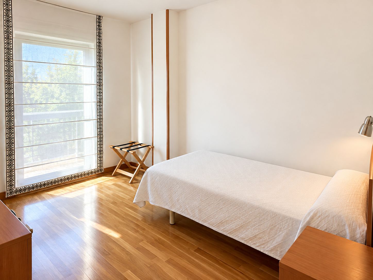 A bedroom with a bed, a nightstand, a lamp, a window with a view, and a wooden floor.
