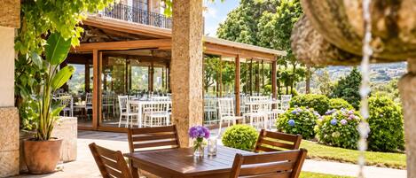 An outdoor patio with wooden tables and chairs, surrounded by greenery and a view of a building with a balcony.
