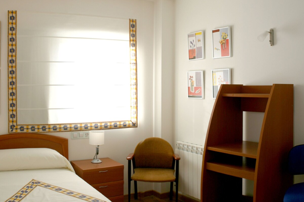 A bedroom with a bed, a chair, a desk, and a window.