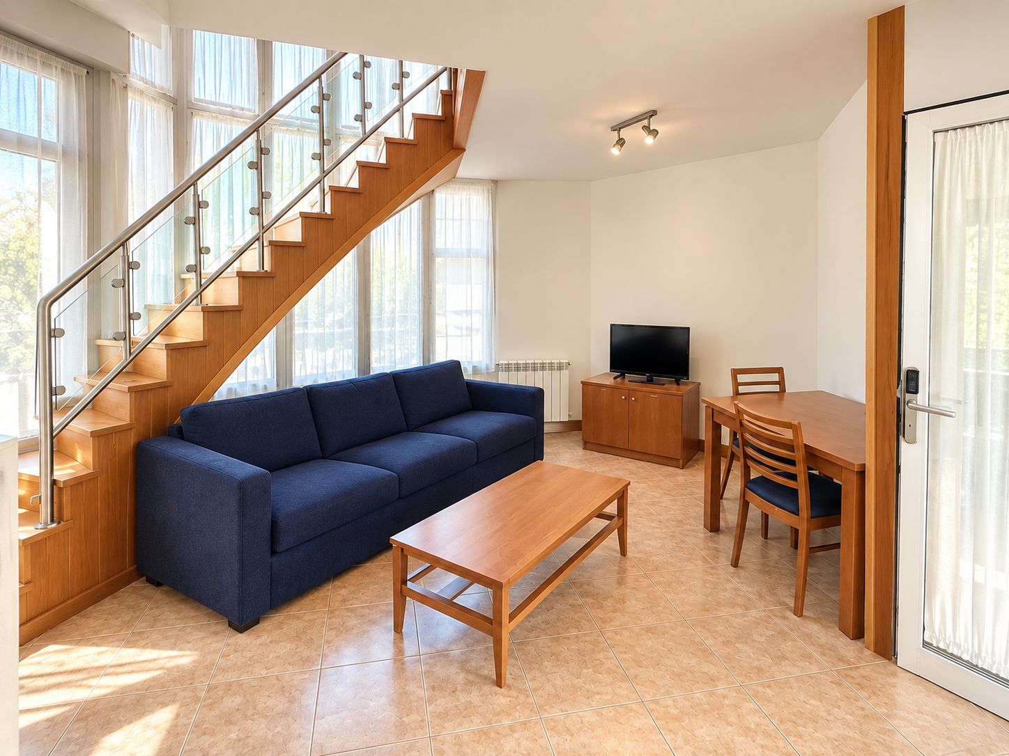 Una sala de estar moderna con una escalera de madera, un sofá azul, una mesa de centro de madera, un televisor en un mueble bajo y un comedor con una mesa y sillas.