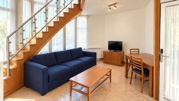 Una sala de estar moderna con una escalera de madera, un sofá azul, una mesa de centro de madera, un televisor en un mueble bajo y un comedor con una mesa y sillas.