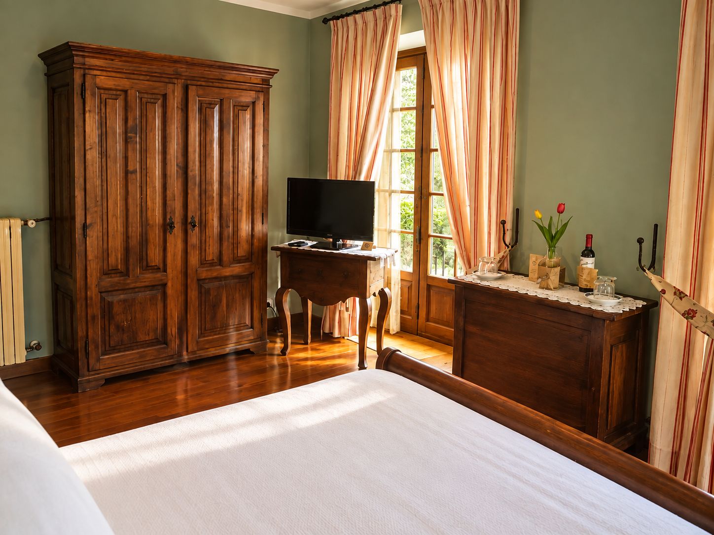 A bedroom with a wooden wardrobe, a bedside table with a television, and a window with curtains.