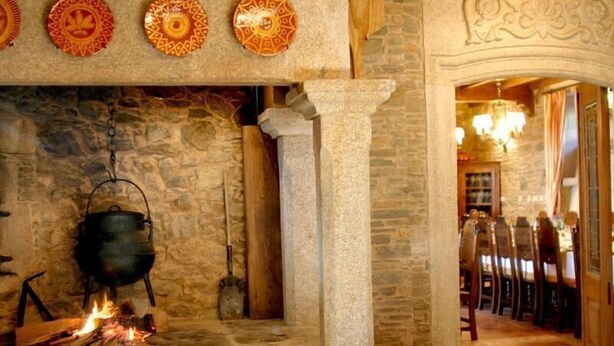 A stone fireplace with a pot on a fire, surrounded by stone walls and a wooden archway leading to a dining area.