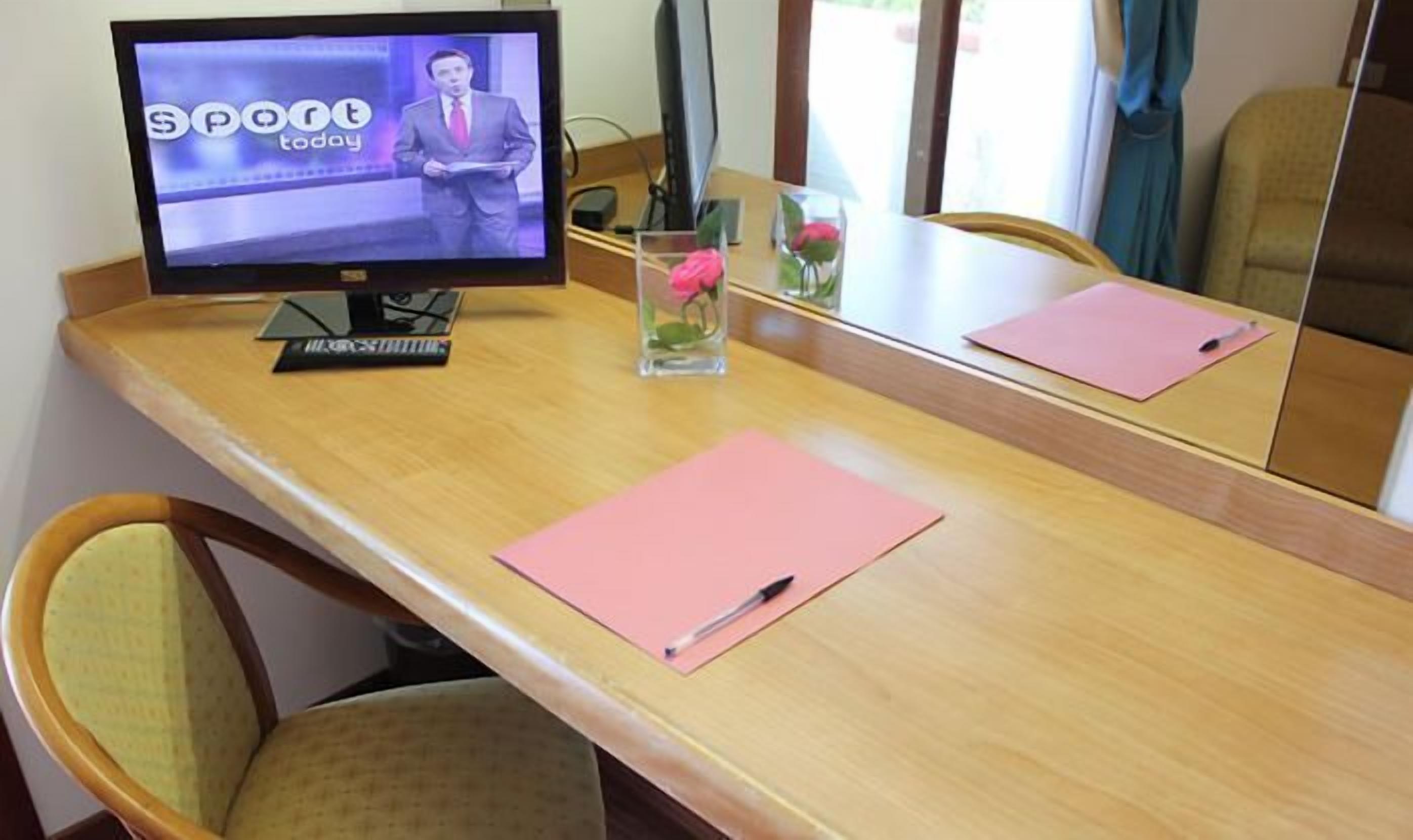 A hotel room with a wooden desk, a television displaying a news channel, a vase with flowers, and a mirror reflecting the room.
