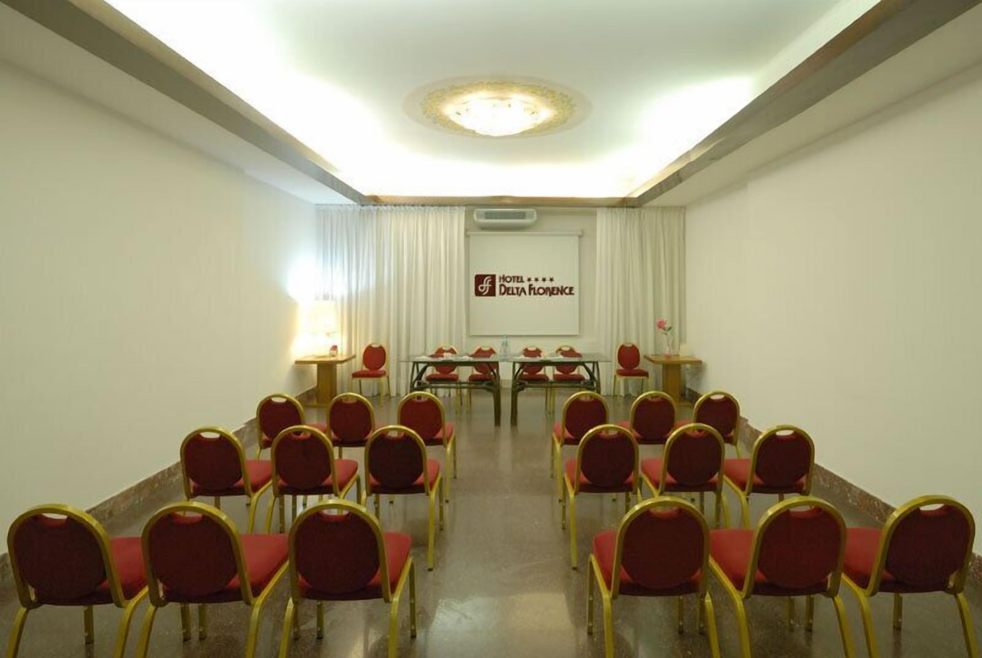 A conference room with rows of red chairs and tables, a ceiling light fixture, and a sign for Hotel Delta Florence.