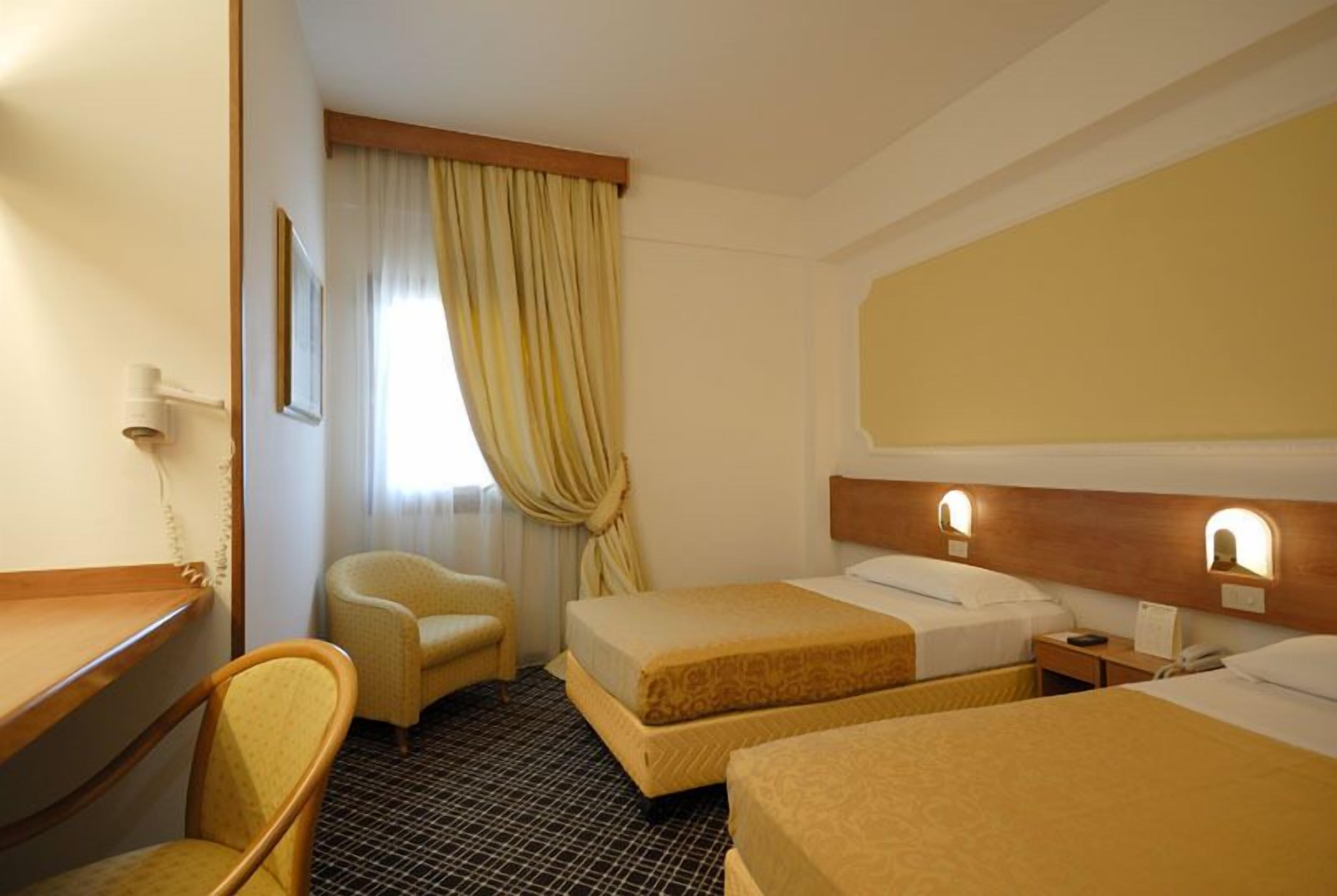 A hotel room with two beds, a desk, a chair, and a window with curtains.